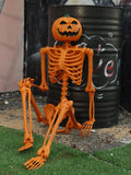 Orange skeleton with a jack-o'-lantern head sitting against a black barrel on grass