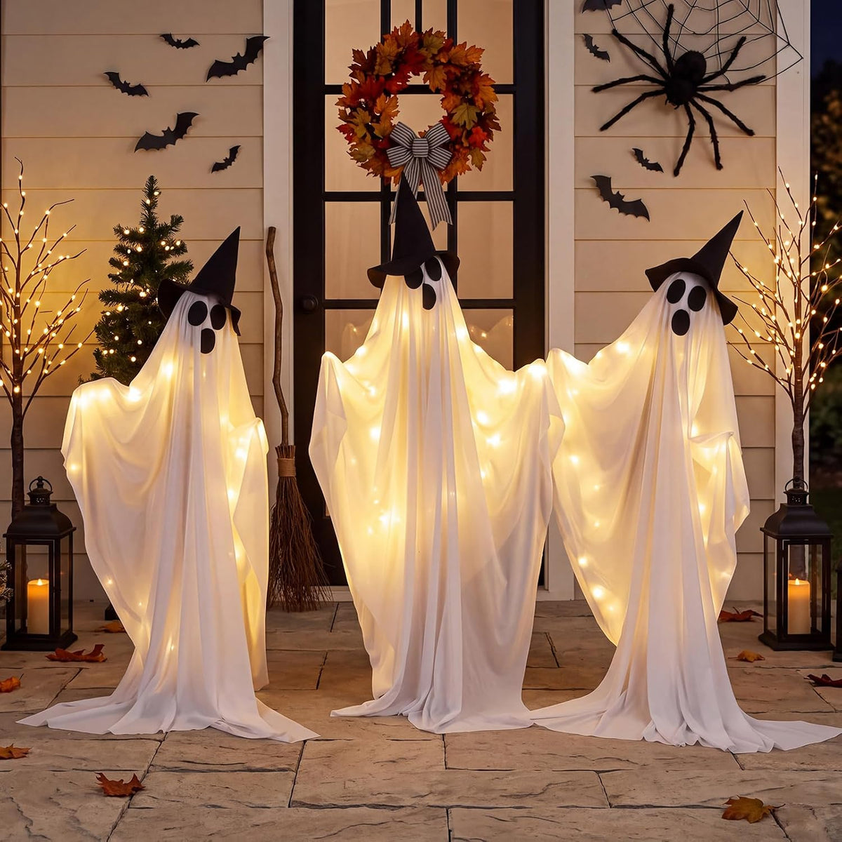 Three glowing white draped ghosts in black witch hats standing on a porch with bats on the wall